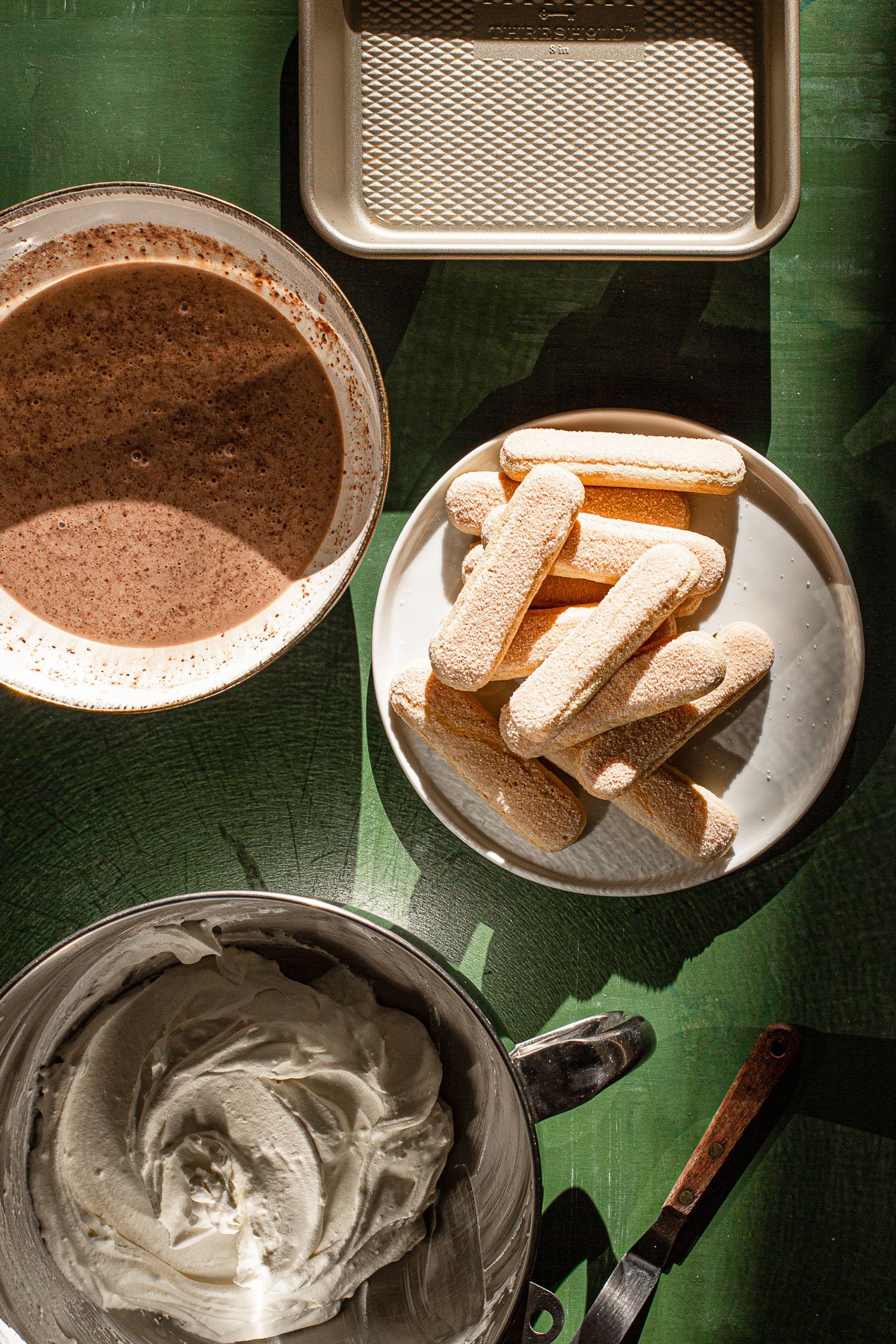 8x8 pan, milo and milk in a bowl, lady fingers on a plate, whipped cream in a mixing bowl.  Spatula on the side on a green backdrop.