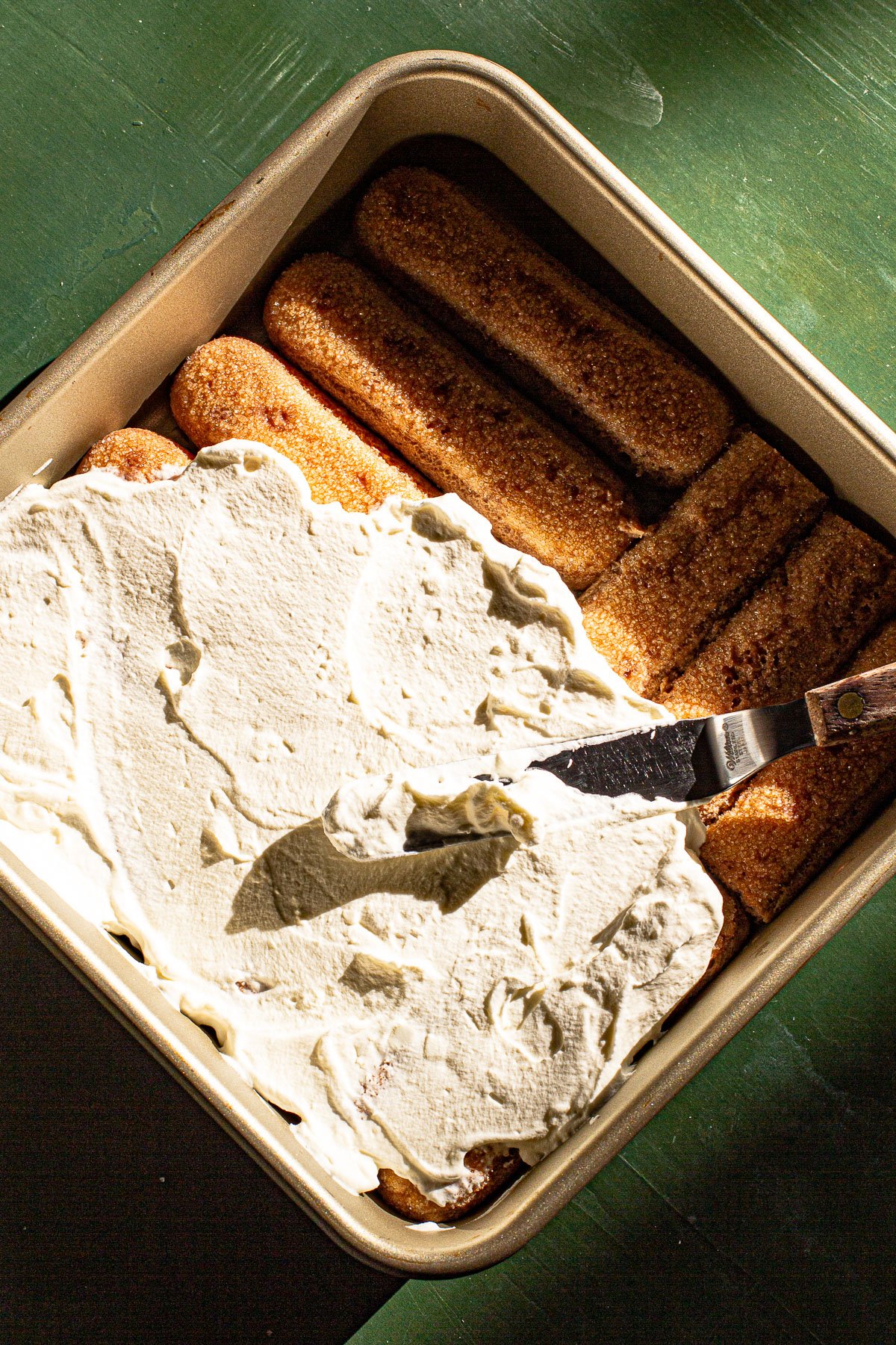 Spreading whipped cream on top of Milo soaked lady fingers using a spatula.  Green Backdrop.