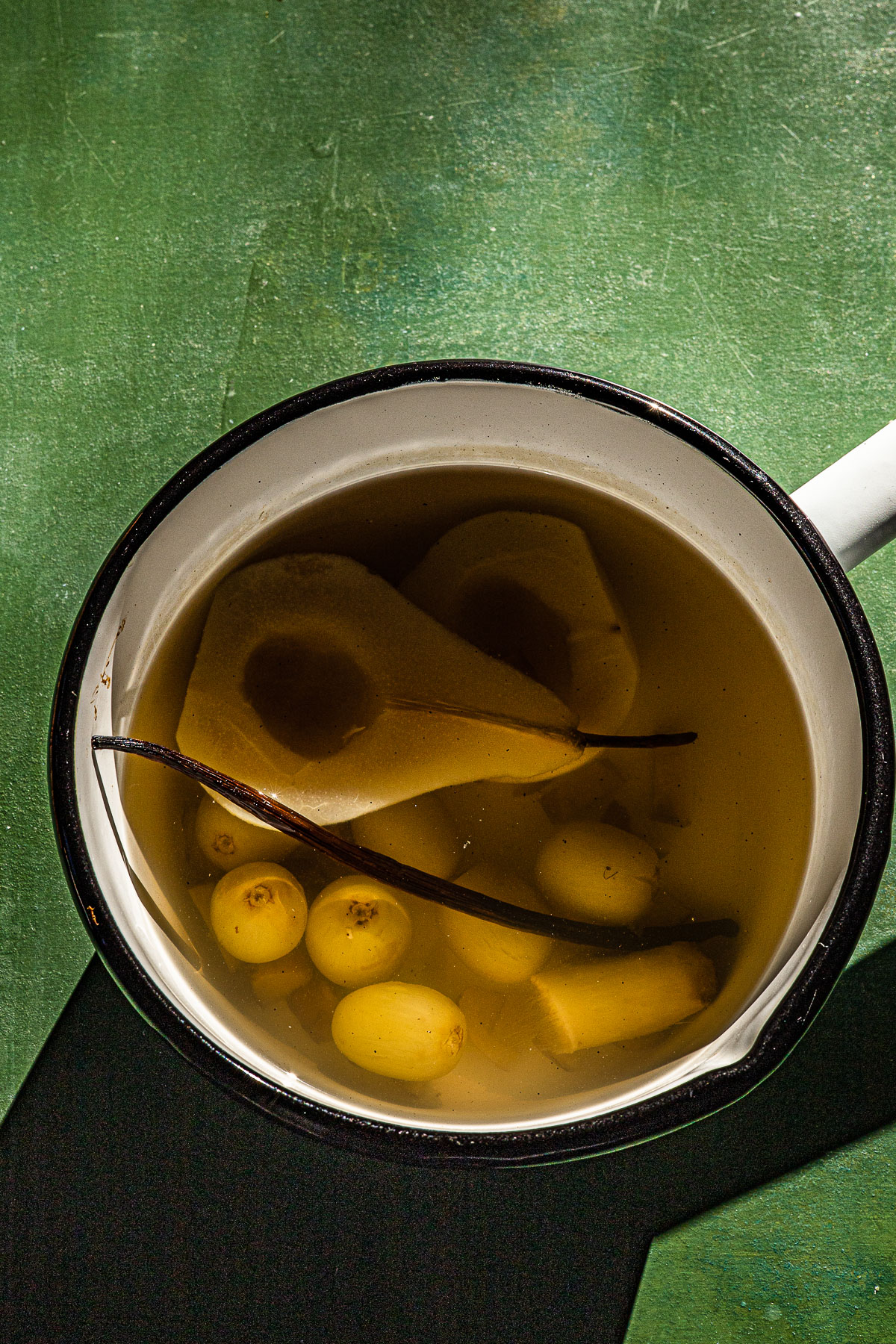 Poached pear and grapes with ginger and vanilla.  In a white enamel pot on a green backdrop.