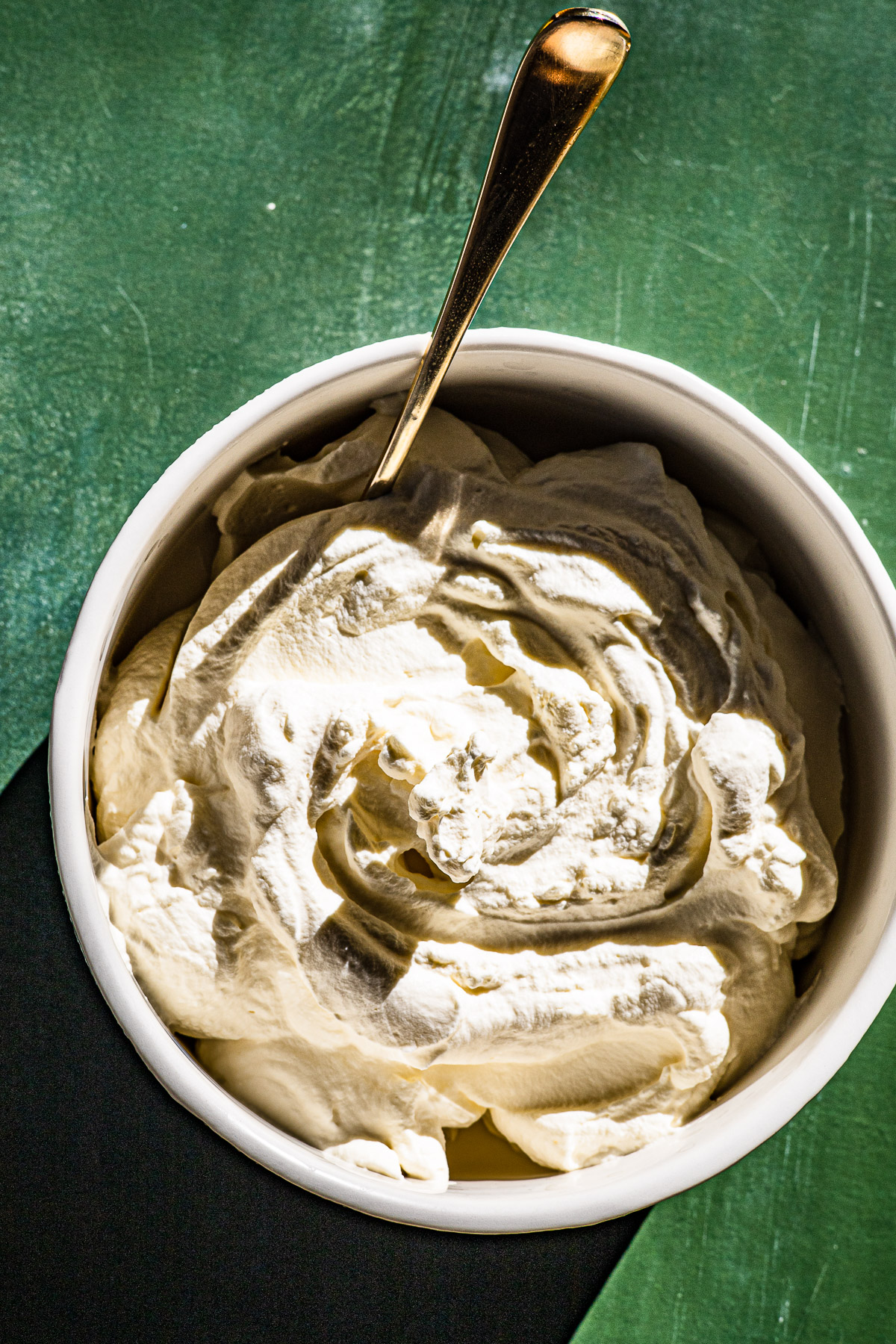 Fresh whipped cream in a bowl with a gold spoon.  On a green backdrop.