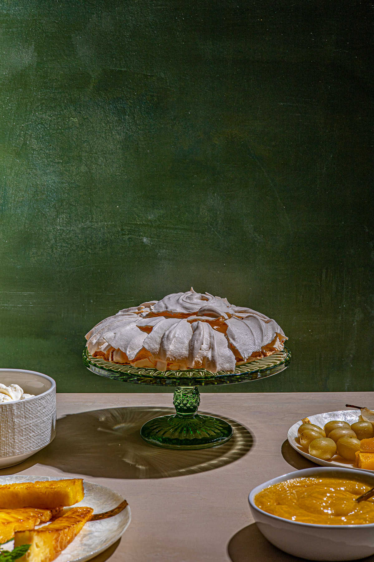 Tropical Pavlova ingredients.  Whipped cream in a bowl, calamansi curd in a bowl, grilled pineapple on a plate.  With a green backdrop.