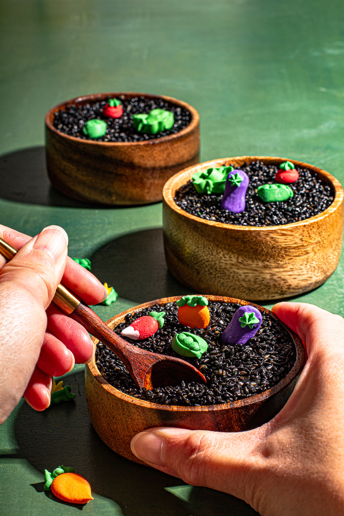 one hand holding a spoon, one hand holding the bowl of black sesame kalamay. With 2 wooden bowls of kalamay in the back on a green backdrop.