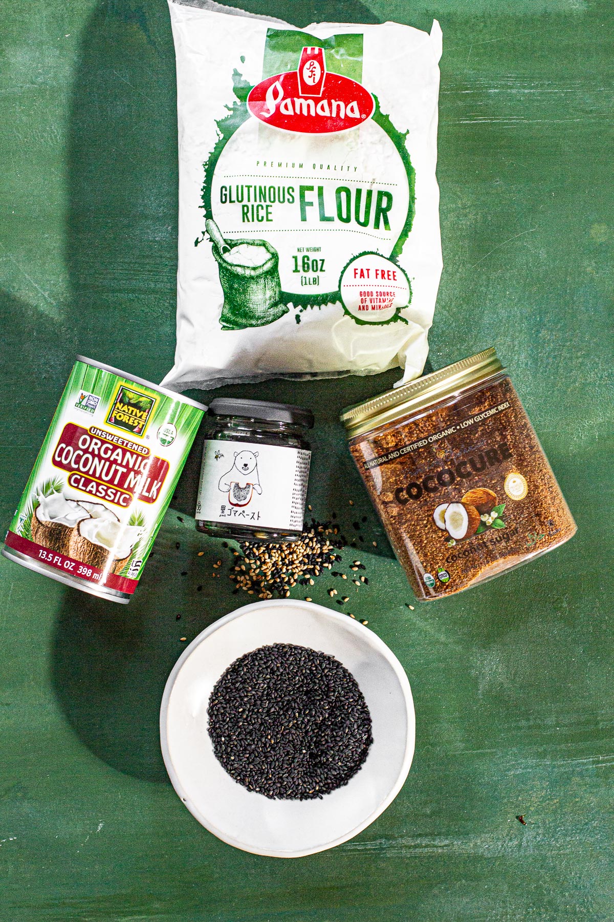 glutinous rice flour in a bag, coconut milk in a can, black sesame paste in a jar, coconut sugar in a jar, black sesame seeds on a plate on a green backround.
