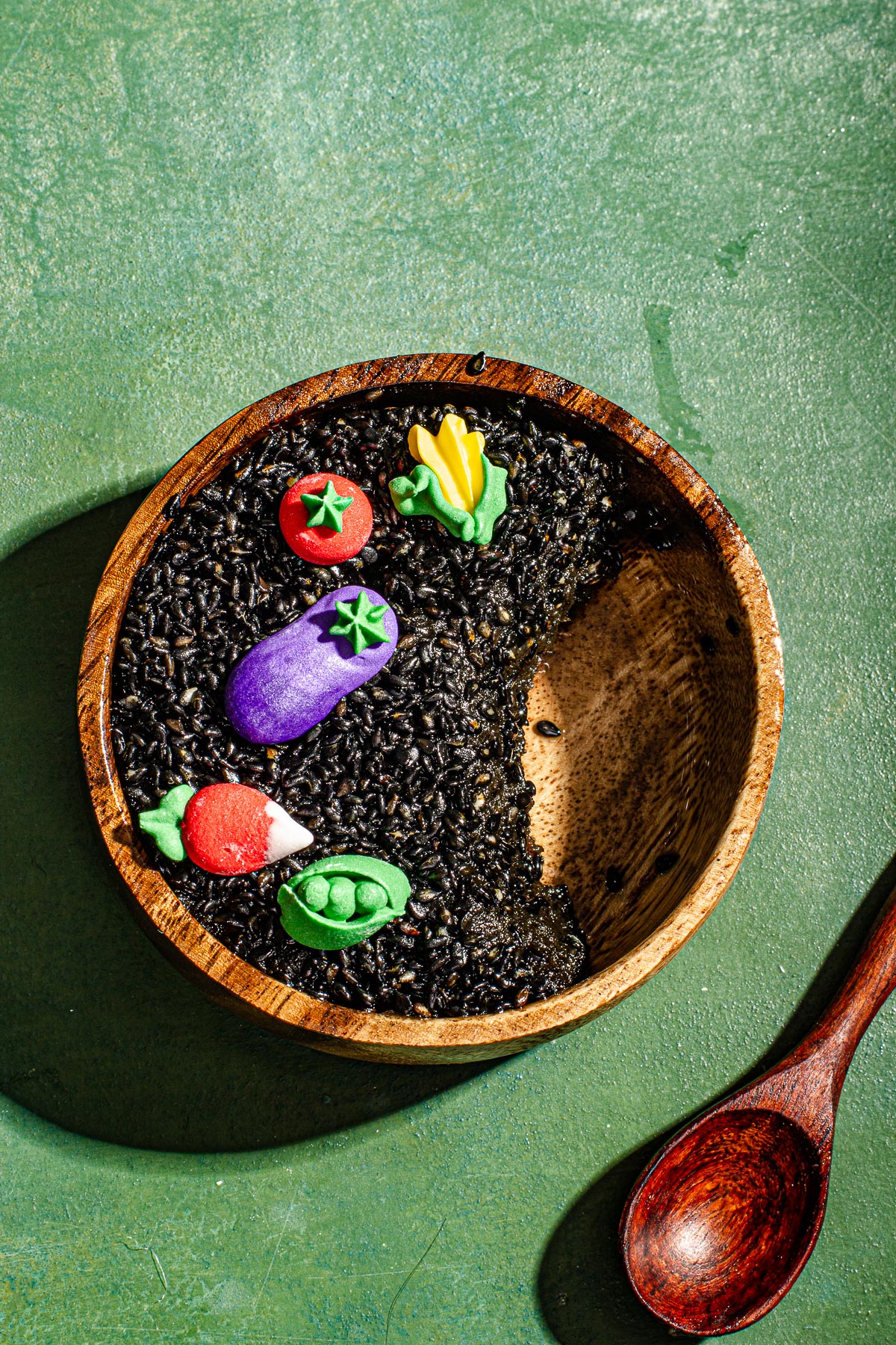 Black sesame Kalamay decorated with sugar vegetables on a green backdrop. With a spoon on the side.