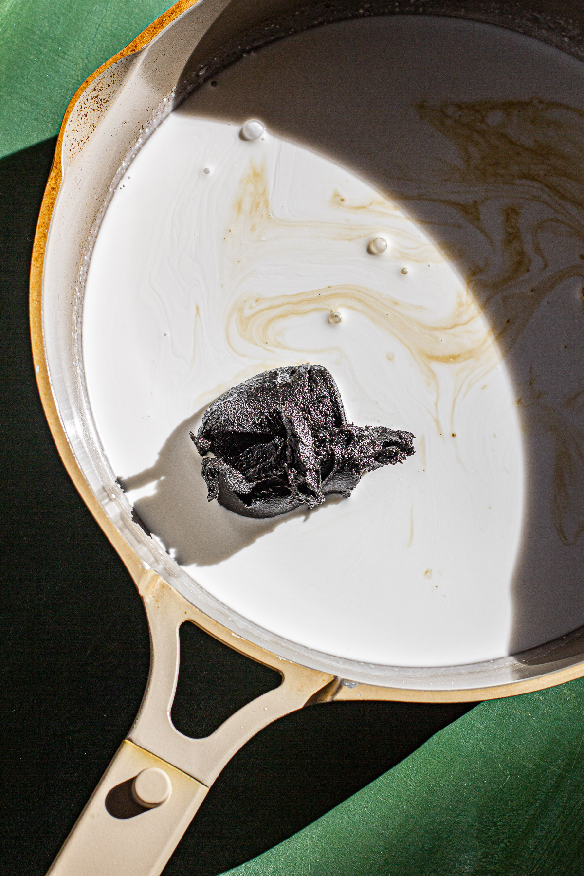 Coconut milk, coconut sugar and black sesame paste in a pan on a green backdrop.