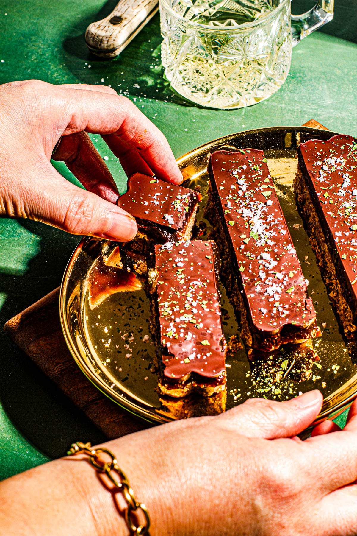 One hand grabbing a Millonariyo Bars (Filipino Style Millionaire Bars) on a gold plate on a small cutting board. All on a green background. With glass of Champane in the back.
