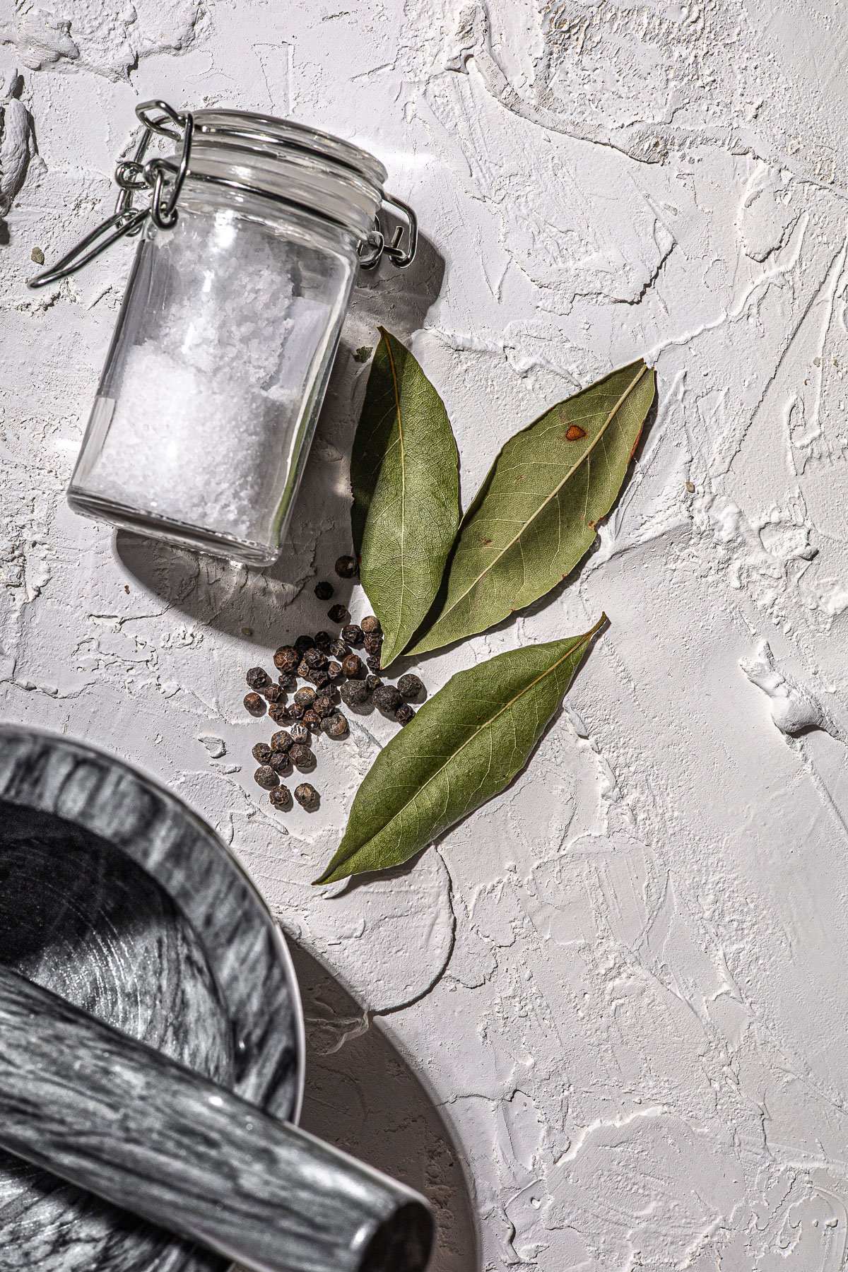 Bottle of Salt, 3 Dried Bay leaves, pepper corns and mortar and pestle on a white background.