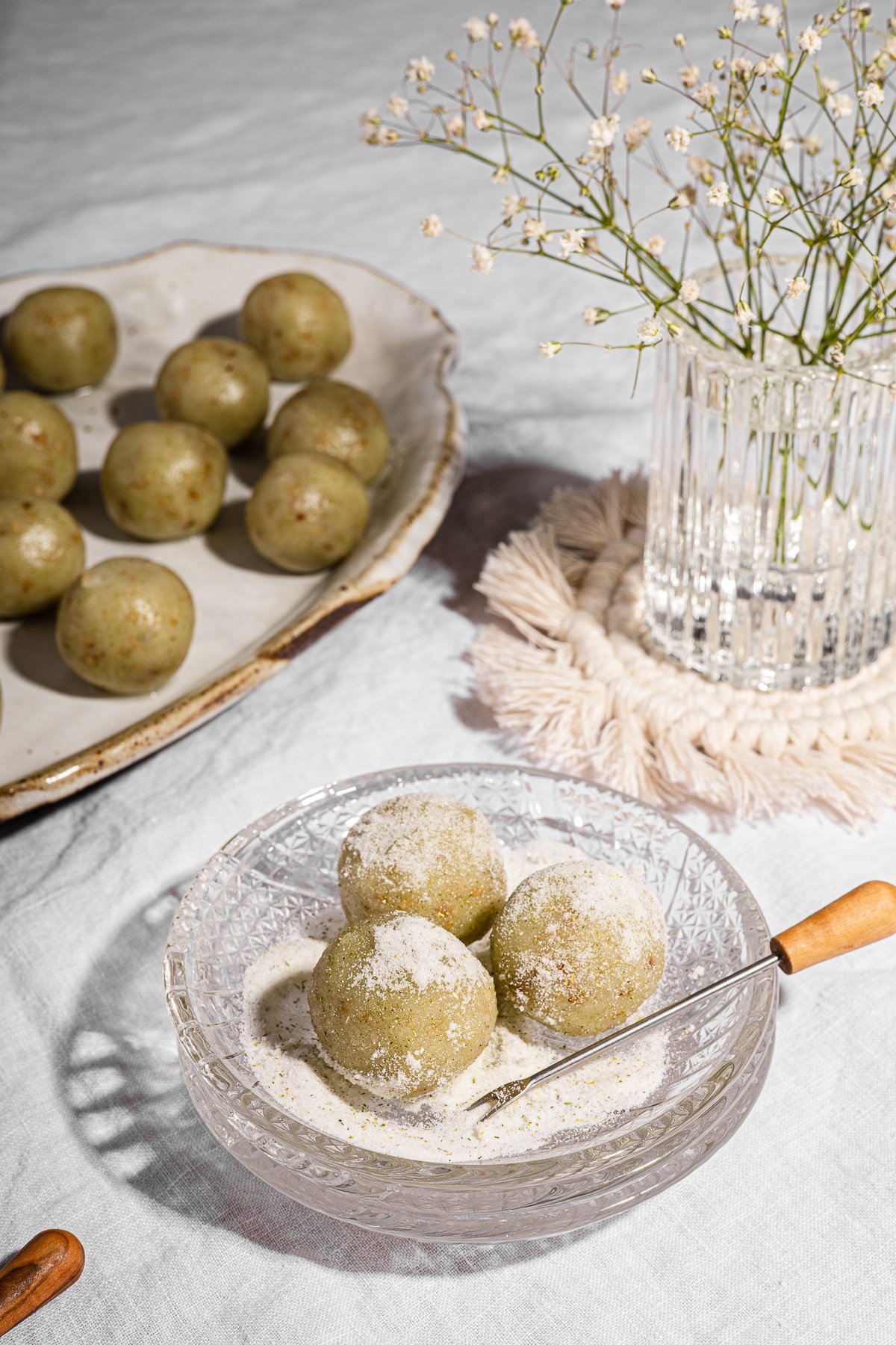3 Balls of Mache (Buko Pandan Mochi Balls) on a glass plate with mini tongs on top of a macrame coaster.  Small vase of baby's breath and mache on a large platter in the background.
