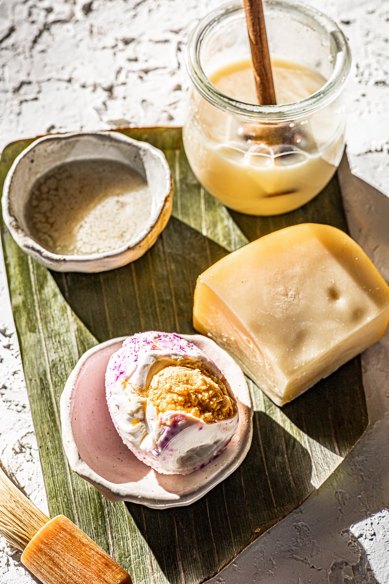 Melted butter in a bowl, condensed milk in a jar with a wooden dripper, itlog na maalat in a bowl and gouda cheese on a banana leaf.