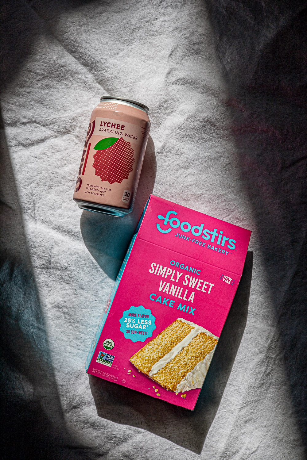 Can of lychee sparkling water and box of vanilla cake mix on a white linen tablecloth.