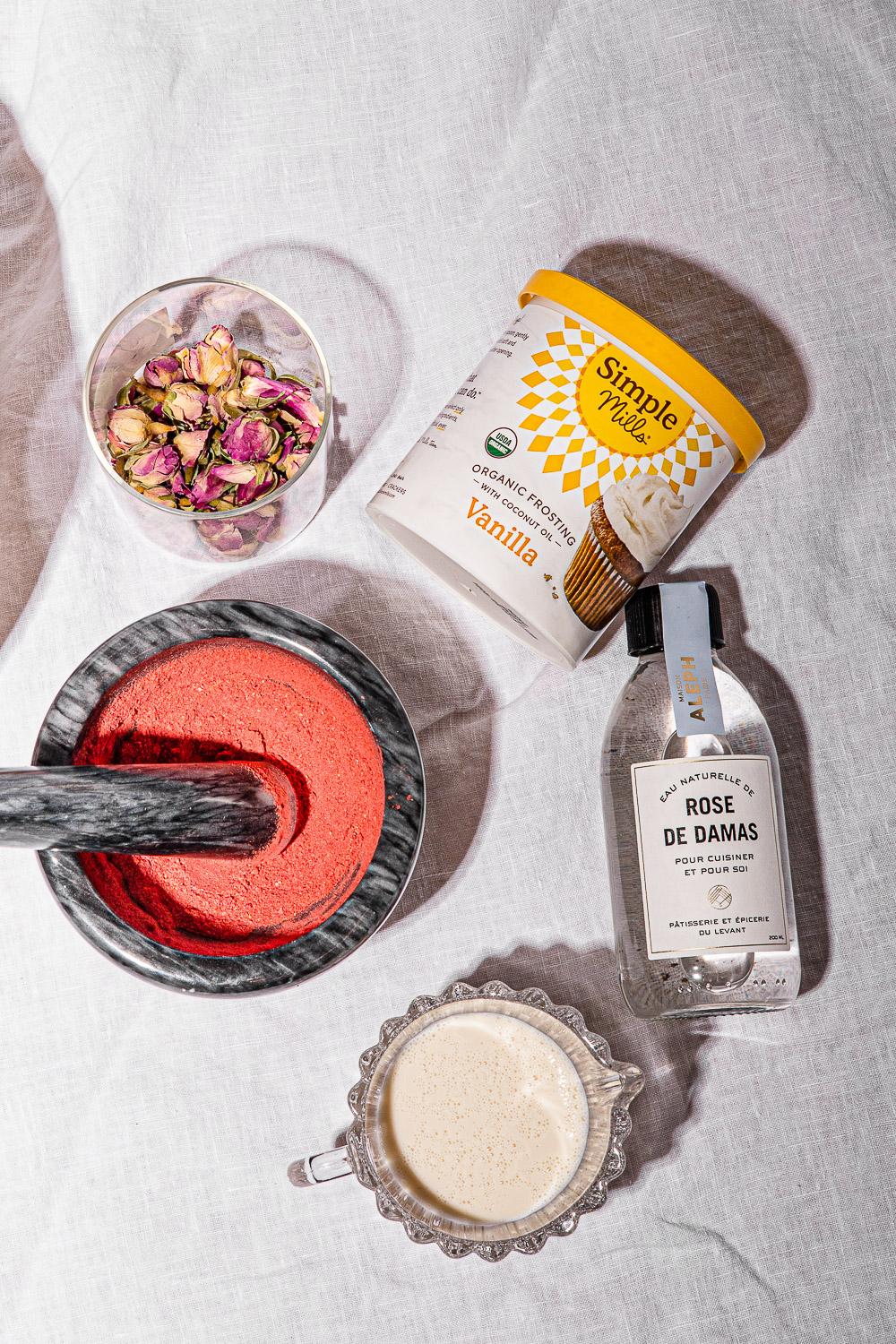 Jar of rose buds, jar of vanilla icing, raspberry powder in grey motar and pestle, bottle of rose water and glass of milk on a white linen table cloth.