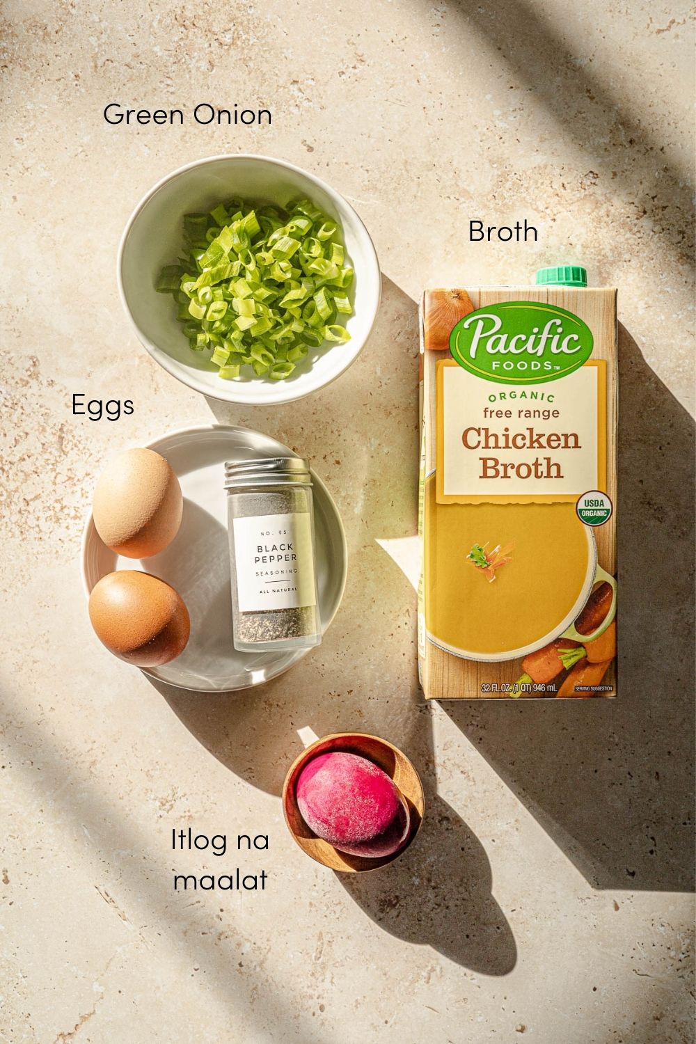 Sliced green onion in a bowl, box of chicken broth, 2 eggs, black pepper in a shaker and a itlog na maalat in a small bowl.