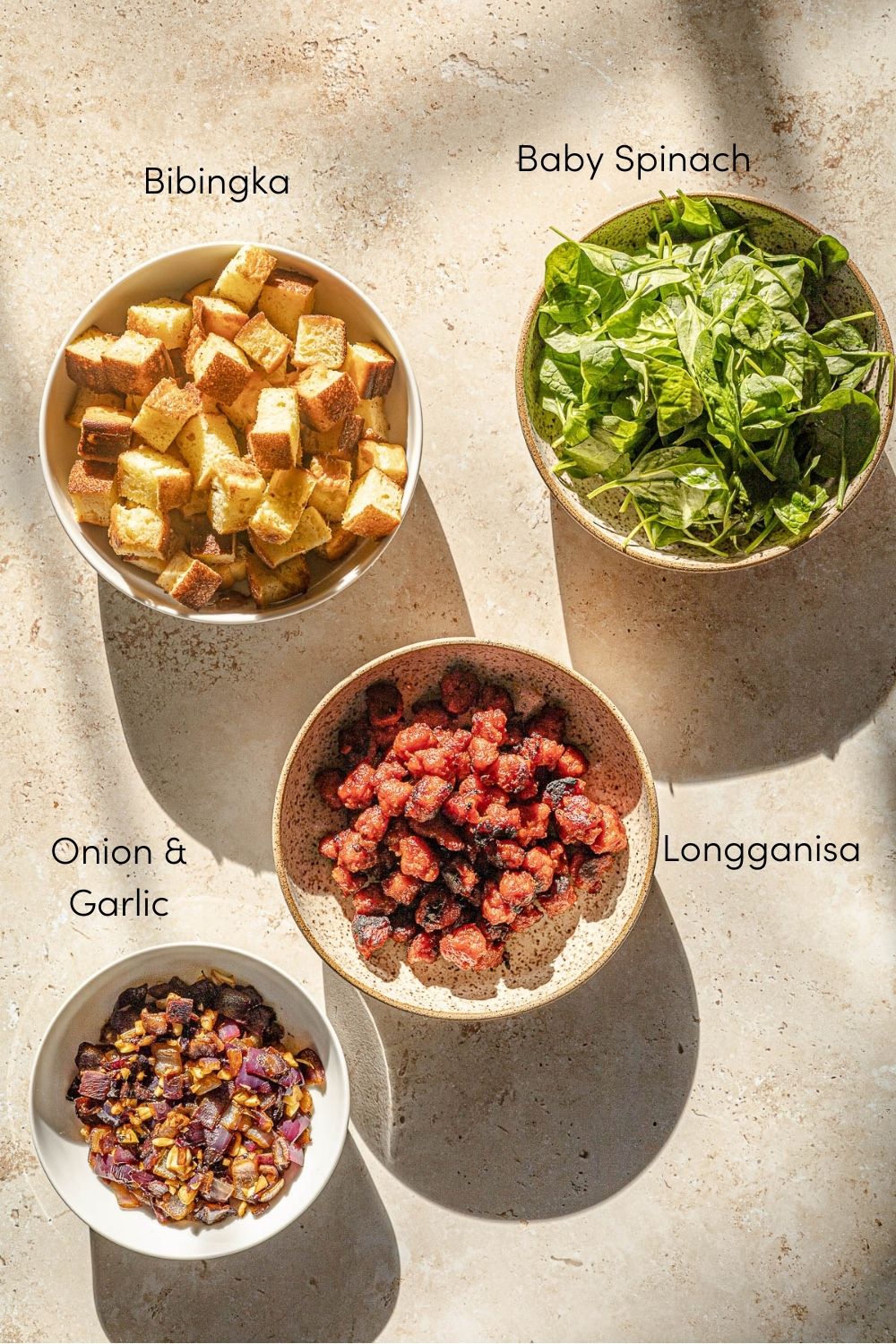 Bibingka in a bowl, baby spinach in a bowl, cooked longganisa in a bowl and cooked onion and garlic in a bowl.