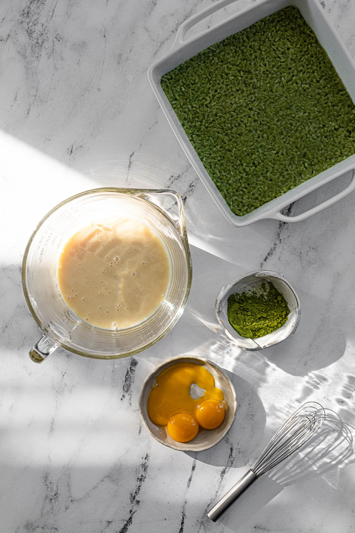Cooked Matcha Biko in a white pan, condensed milk in a glass bowl, matcha on plate, egg yolks in a plate.  Whisk on the side.