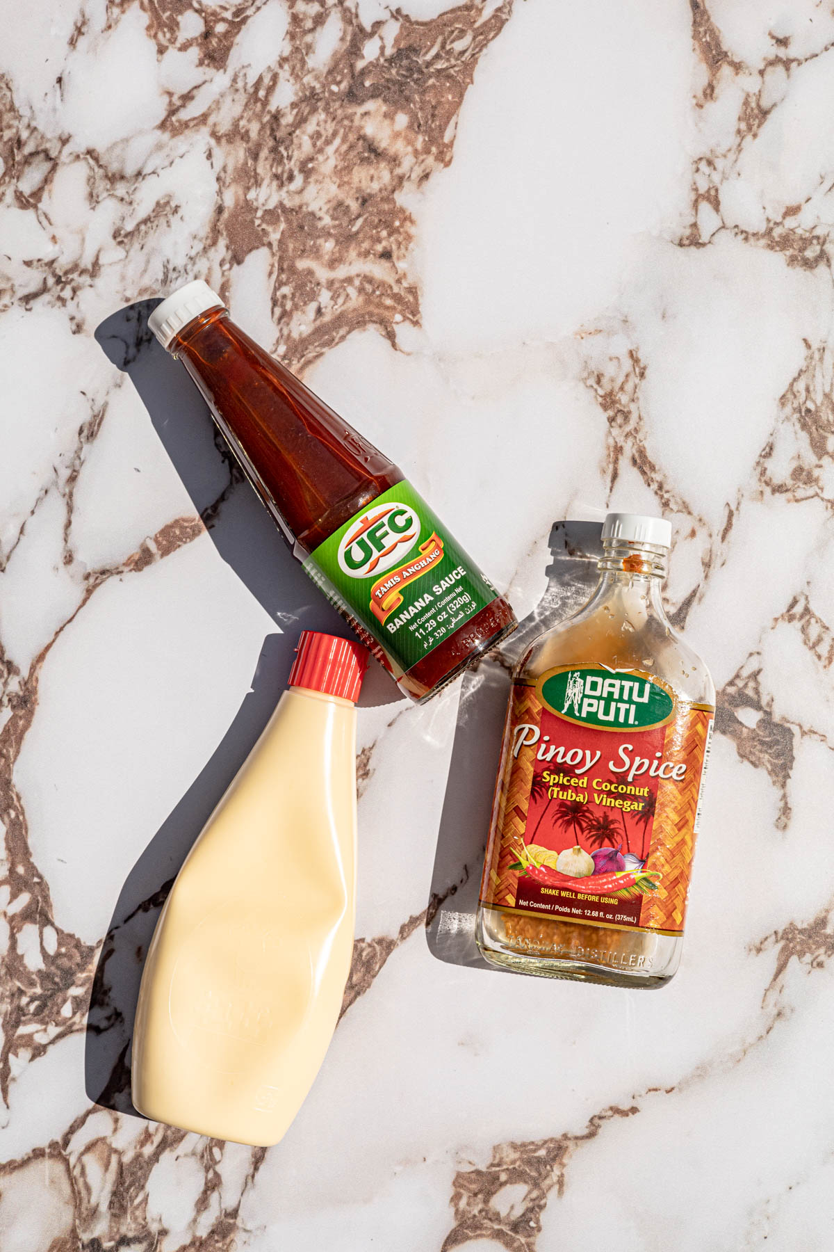 Bottle of banana ketchup, bottle of vinegar and bottle of kewpie mayonnaise on a marble countertop.