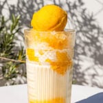 A glass of Mango Halo Halo, topped with a scoop of mango sorbet, sits on a white surface with plant shadows in the background.