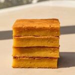 Four rectangular pieces of cornbread are stacked on a light-colored surface, reminiscent of the golden hues found in Peach Mango Bibingka, with sunlight casting gentle shadows in the background.